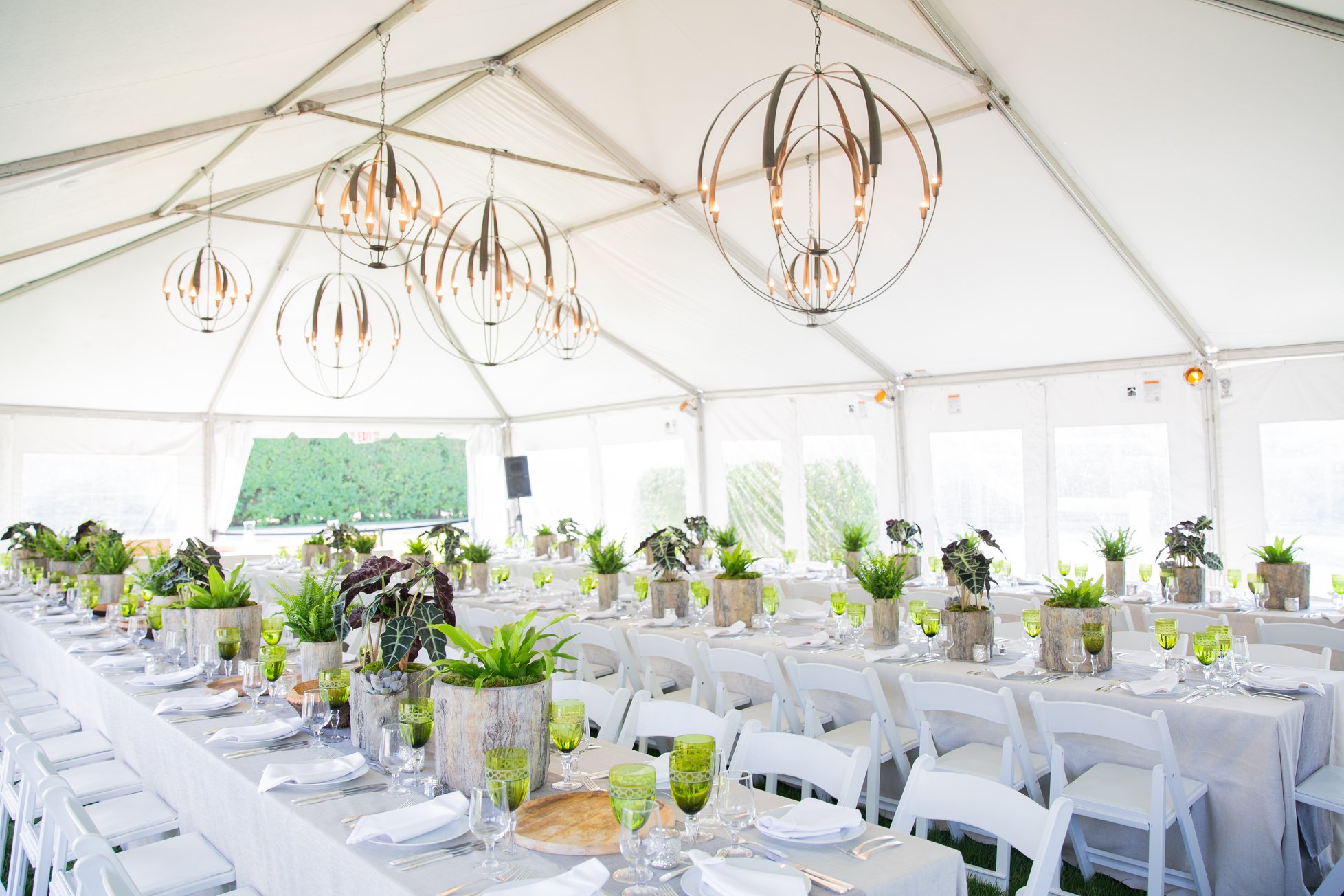 A white event tent in the Hamptons set for dinner, with spherical chandeliers, long tables, green glassware, and potted-fern centerpieces