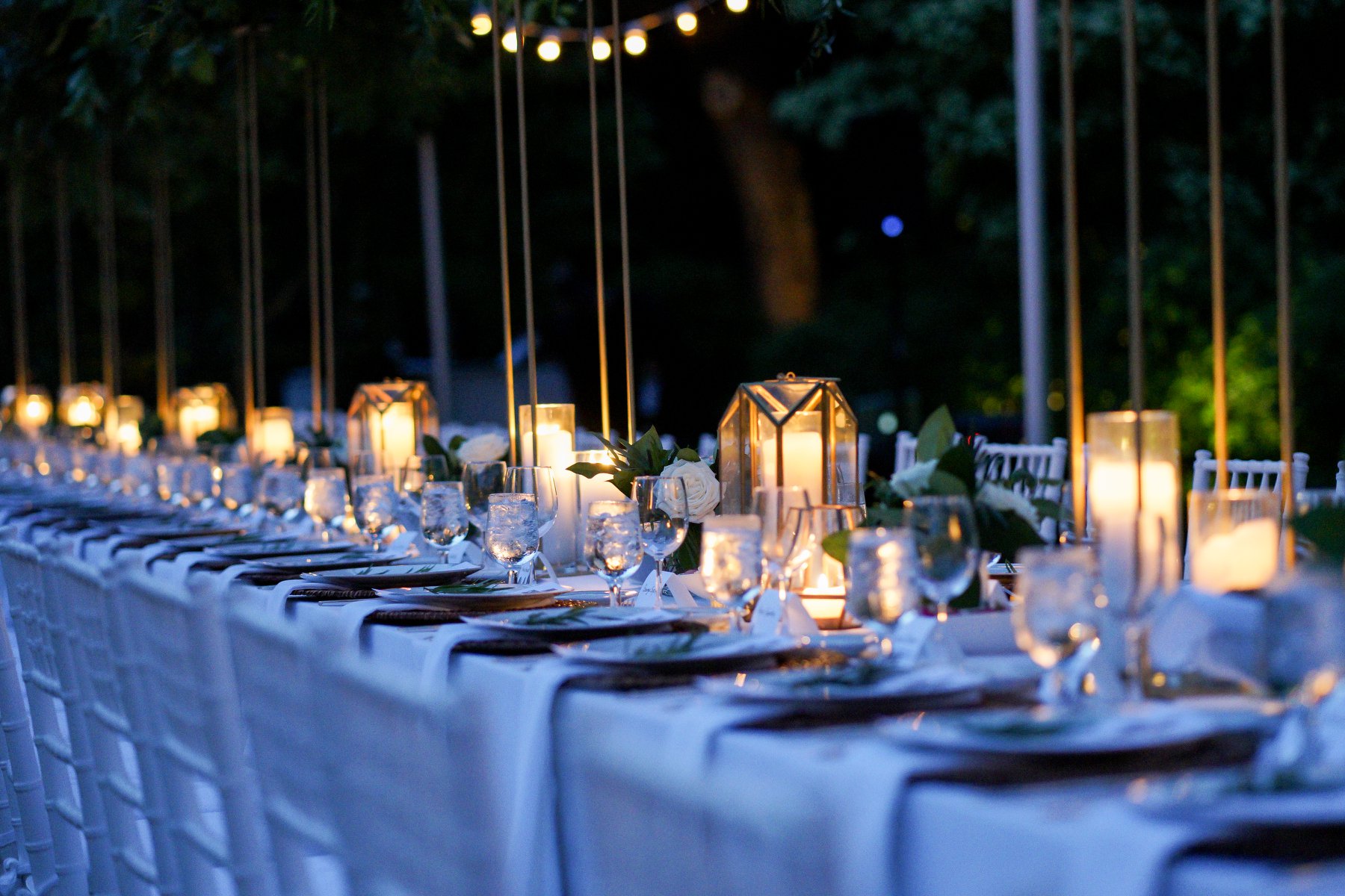 An outdoor Hamptons dinner table at dusk lit by candles and hurricane lanterns, with white roses and string lights overhead