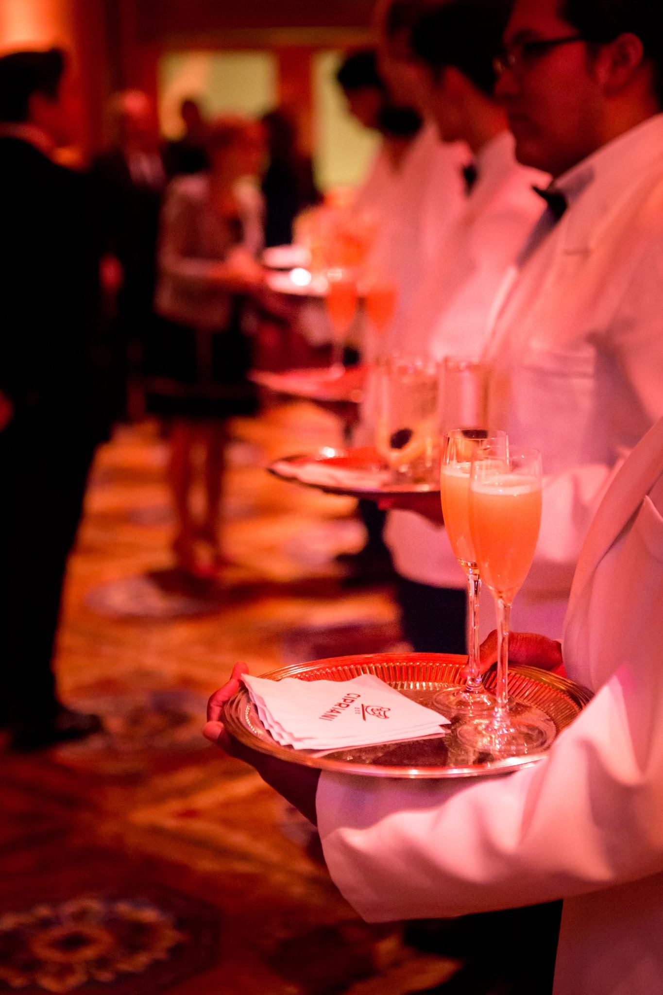 A line of white-jacketed servers passing trays of champagne cocktails in a warm, red-lit ballroom at a New York City gala
