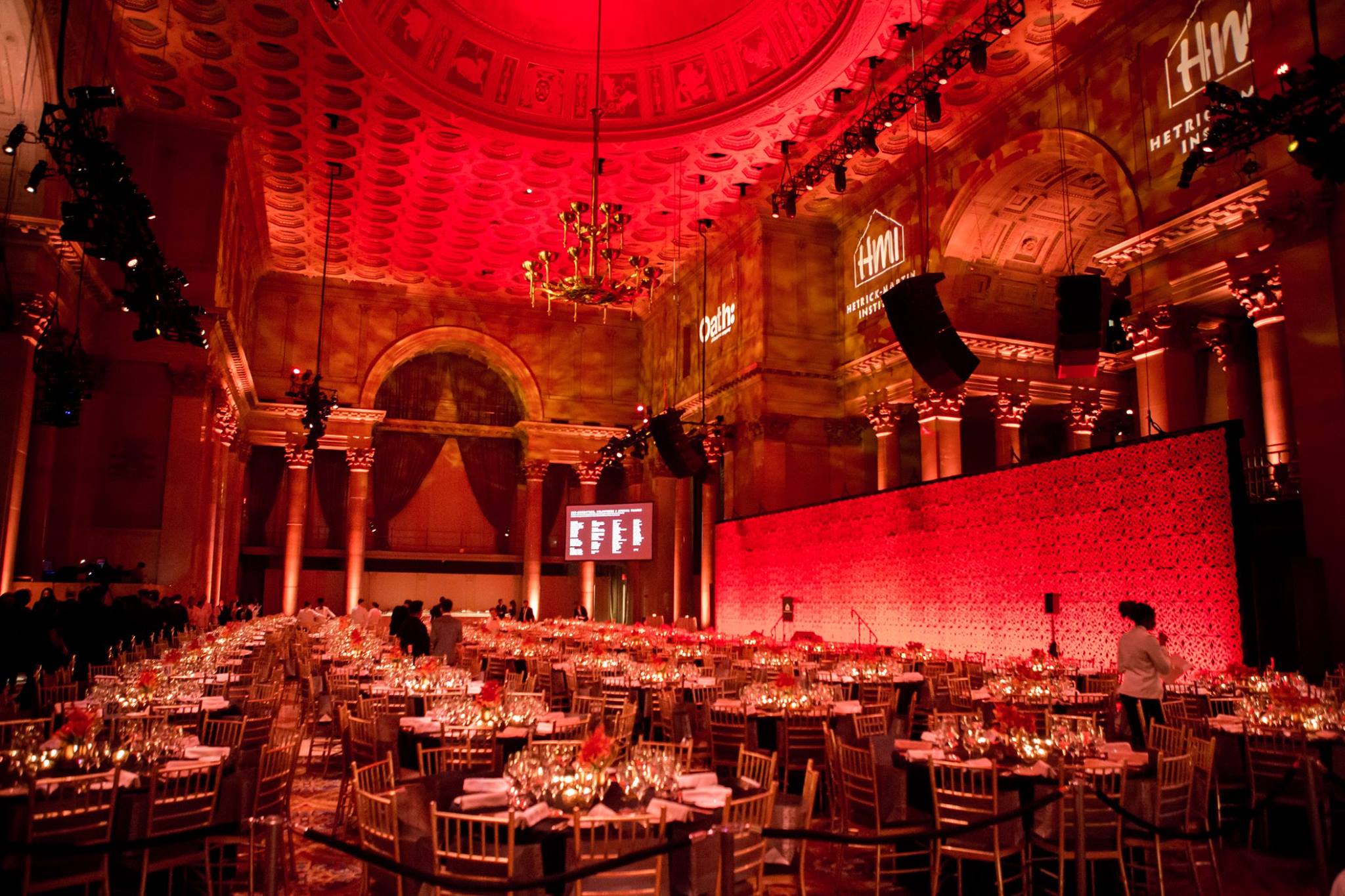 A grand landmark ballroom bathed in red light, with gilded columns, a domed ceiling, and round tables set for a New York City gala