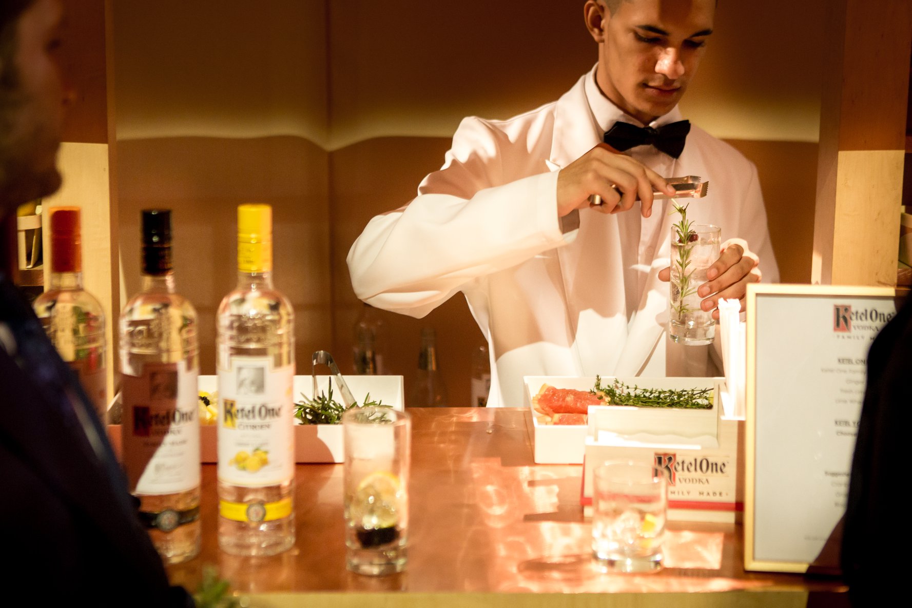 A bartender in a white dinner jacket garnishing a cocktail at a copper-topped bar during a New York City gala